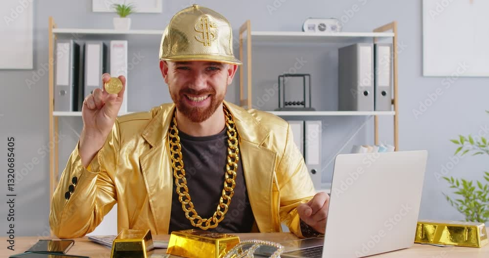 Portrait of happy smiling funny young man stock trader wearing gold suit holding bitcoin in hands and looking cheerful at camera sitting at the desk at home or at office with laptop. 4k video.