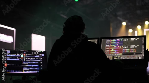 A stage lighting technician operates a control panel during a live concert, surrounded by green stage lights. A DJ performs in the background.