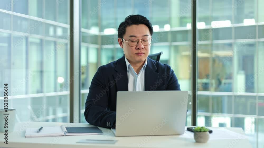 Focused Korean businessman working on a laptop in a modern office with large windows. He thinks about a new project. Focused Korean businessman working on a laptop in a modern office with large windows. He thinks about a new project.