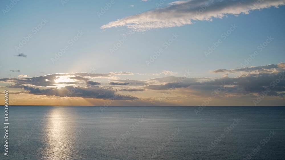 Fototapeta premium Sun shining through clouds over calm ocean at sunset