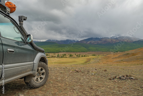 An off-road car for family trips on mud tires stands on an autumn yellow meadow overlooking a green coniferous forest and mountains covered with snow.