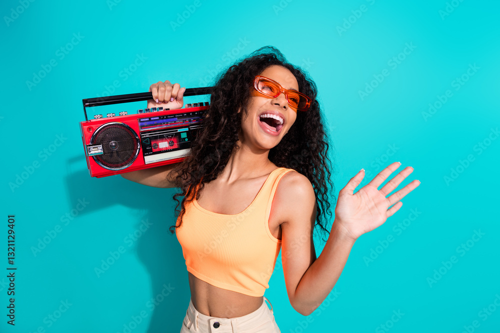 Obraz premium Joyful young woman holding a vintage red boombox, wearing stylish orange sunglasses and top, against a vibrant teal background