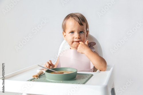 toddler with light brown hair sits in high chair, eating soup bread while wearing pink bib. child looks curiously at the camera, with one finger in their mouth. Concept of childhood and healthy eating photo