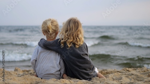 Blonde sisters sitting closely on sandy shore, embracing while gazing at ocean waves during peaceful evening light, sharing quiet moment of childhood connection and joy