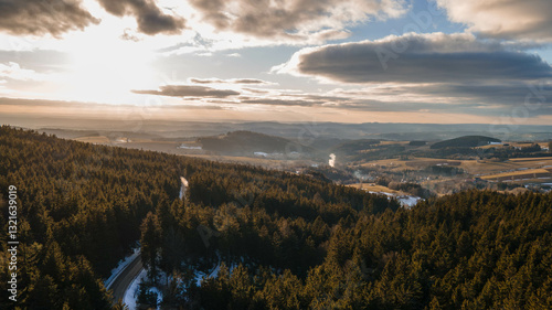 A stunning aerial view of Červenovodské Sedlo in Orlické Hory, Czech Republic, bathed in golden sunset light. Dense evergreen forests contrast with fields, creating a breathtaking rural landscape.