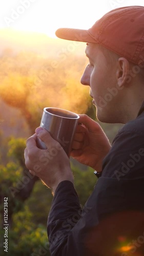 Vertical screen, male traveler wearing bright orange cap savoring steaming hot beverage in metal mug during golden sunrise, surrounded by lush green forest and mountain landscape