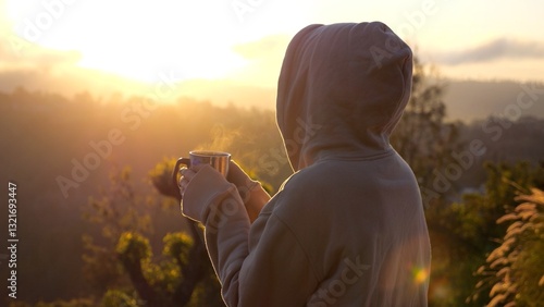 Peaceful woman in hooded sweatshirt standing with steaming mug, enjoying golden sunrise illuminating scenic mountain valley landscape with soft morning light and warm colors