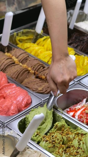 Vertical screen, ice cream vendor serving a scoop of pistachio ice cream, adding chopped pistachios on top, in a gelateria with many flavors