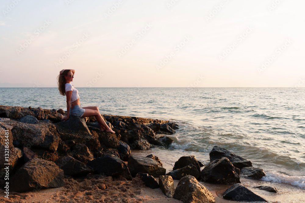 Young woman enjoys the tranquility of the rocky beach while watching the vibrant sunset unfold before her eyes, lost in thought and serenity Young woman enjoys the tranquility of the rocky beach while watching the vibrant sunset unfold before her eyes, lost in thought and serenity