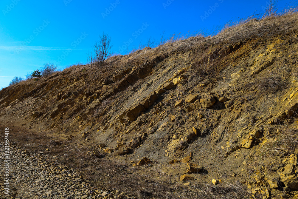 The stones have yellow and brown hues, and layers of different rocks are visible. Dry grass and sparse bushes grow on the top of the hill. The sky is blue, cloudless. The shot was taken on a sunny day The stones have yellow and brown hues, and layers of different rocks are visible. Dry grass and sparse bushes grow on the top of the hill. The sky is blue, cloudless. The shot was taken on a sunny day