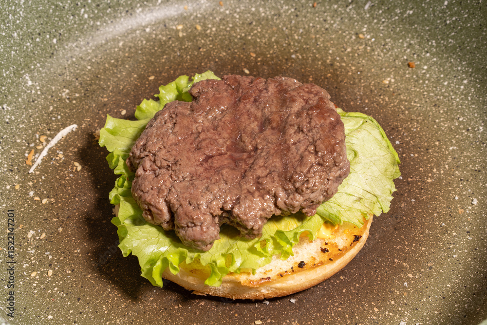 A cooked hamburger patty atop fresh lettuce on a toasted bun in a frying pan, capturing a rustic stage of homemade burger preparation.