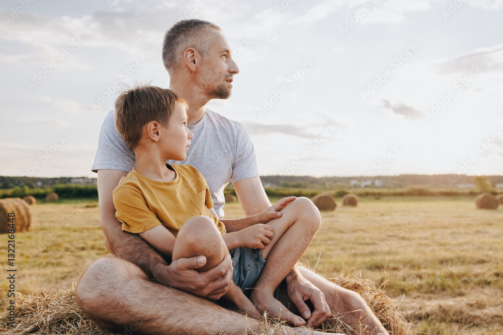 Father and son sitting together on a haystack in a sunlit field, looking into the distance Father and son sitting together on a haystack in a sunlit field, looking into the distance