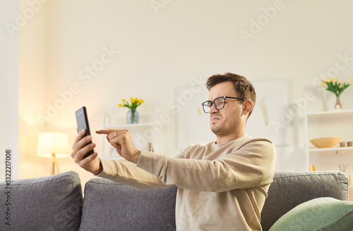 Fényképezés Serious man in glasses sitting on couch at home, holding smartphone at arm length, trying to discern
