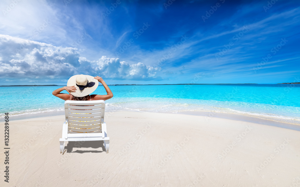 A holiday woman with a sunhat sits on a lounge chair at a tropical paradise beach with turquoise ocean
