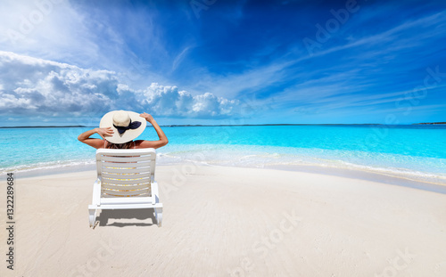 Obraz na płótnie A holiday woman with a sunhat sits on a lounge chair at a tropical paradise beach with turquoise ocean w salonie