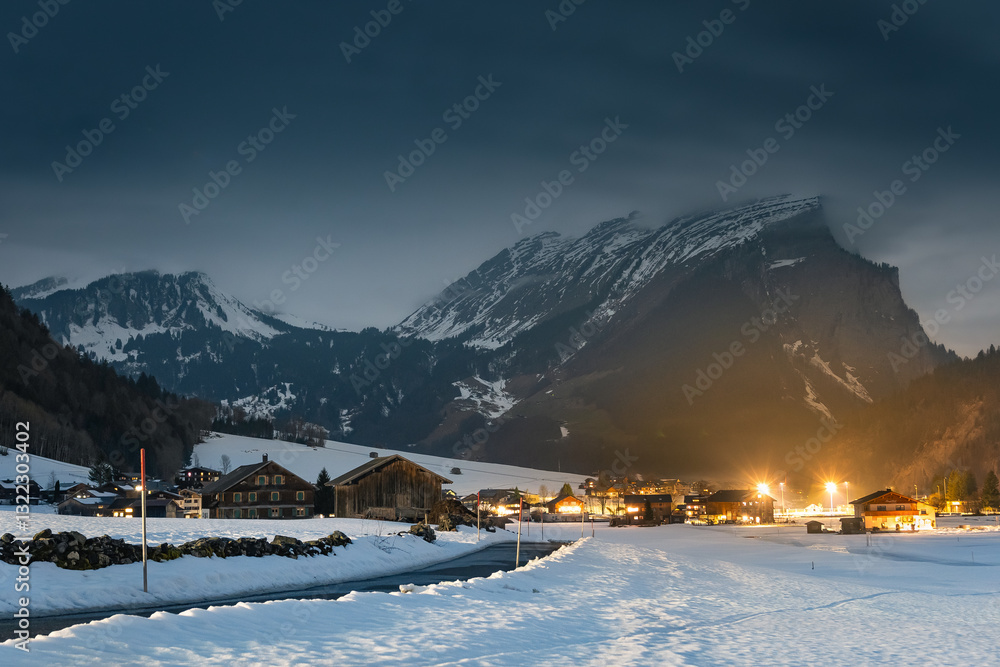 Winter night landscape in the mountains, snow-covered foothills with a small village in the Alps, houses illuminated at night, view from a hiking trail.