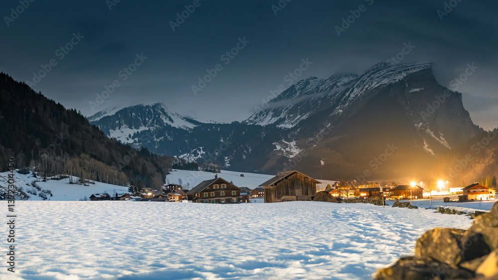 Winter night landscape in the mountains, snow-covered foothills with a small village in the Alps, houses illuminated at night, view from a hiking trail.