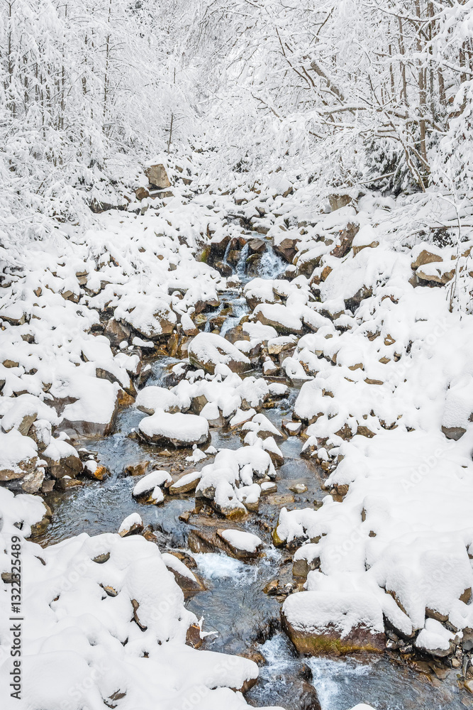 Mountain river in winter, water flows down the riverbed over snow-covered rocks. Winter landscape on a mountain hiking trail.