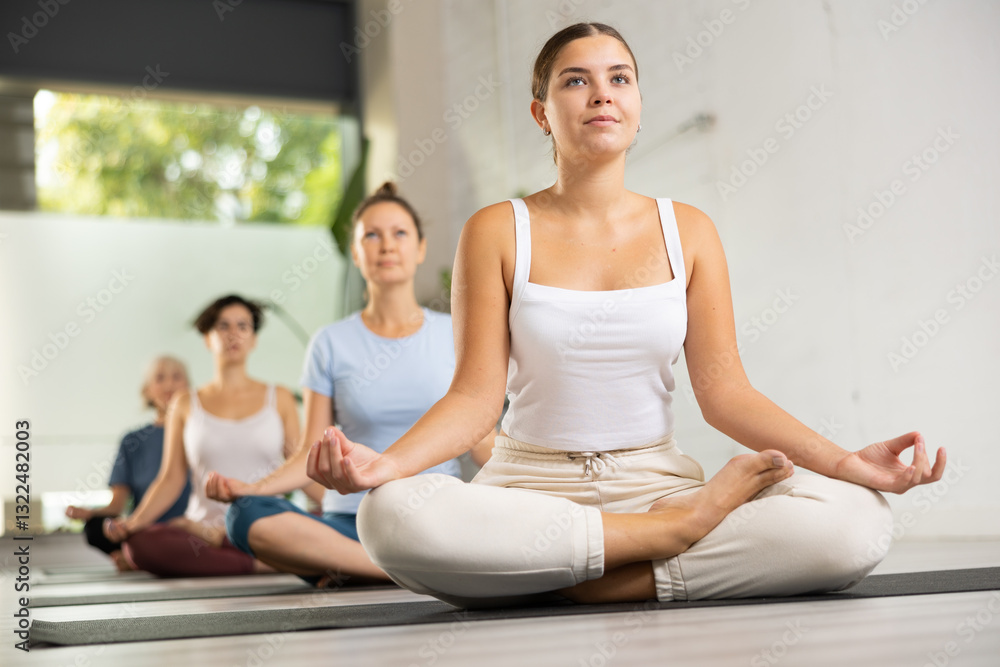 Relaxed young European female sitting in meditation Lotus Pose Padmasana during yoga class with other women Relaxed young European female sitting in meditation Lotus Pose Padmasana during yoga class with other women