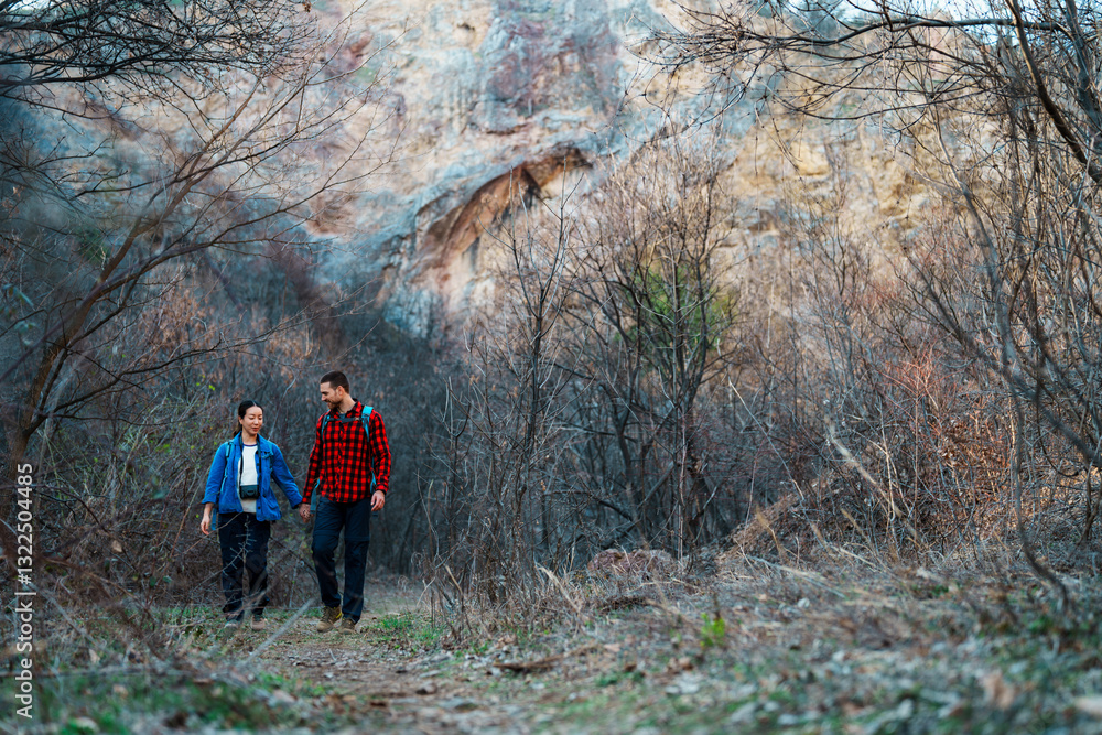 Obraz premium Young couple holding hands while hiking along a scenic trail near a rocky cliff in a bare forest, embracing the cool air of a tranquil autumn day filled with adventure