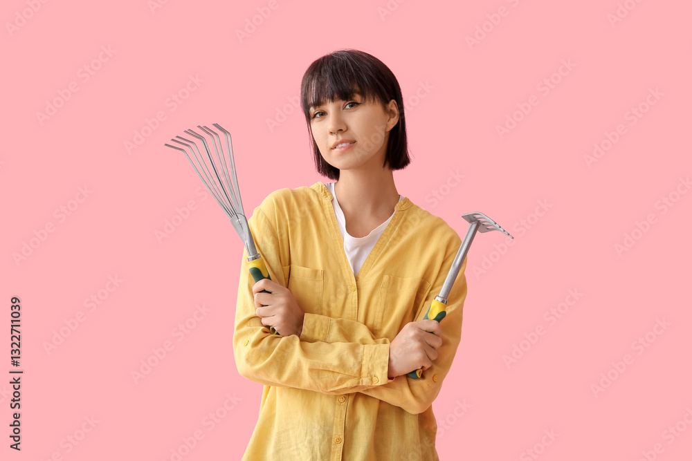 Beautiful young female gardener with gardening rakes on pink background Beautiful young female gardener with gardening rakes on pink background