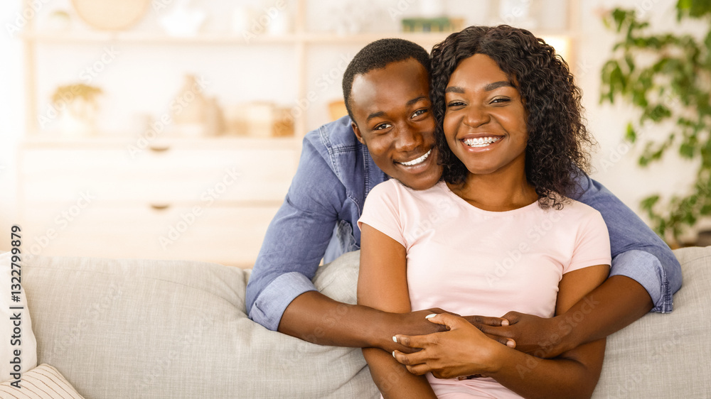 Obraz premium Cheerful african man hugging his smiling wife from behind, spending time together at home, empty space