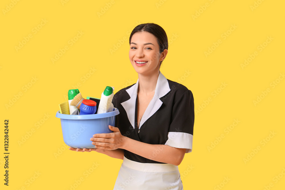 Beautiful young chambermaid holding basin with cleaning products and tools on yellow background Beautiful young chambermaid holding basin with cleaning products and tools on yellow background