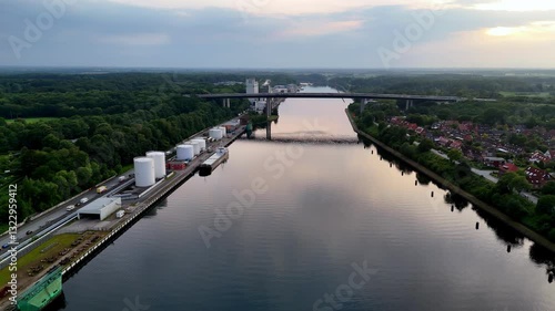 Wallpaper Mural Kiel Canal, evening twilight, aerial view with wide-angle lens, showing the straight waterway reflecting the sky, flanked by dense green forests, industrial tanks, a bridge, and residential houses Torontodigital.ca