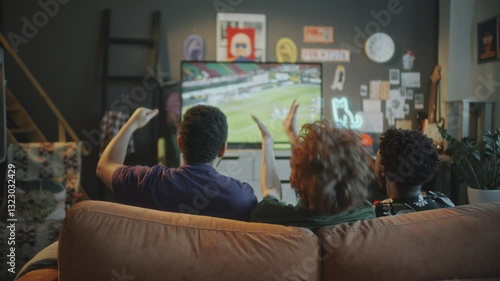 Three young men sitting on couch, raising hands and cheering while watching soccer match on TV in stylish living room with modern decor. Rear view