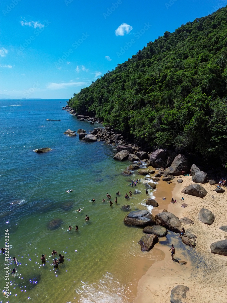 Obraz premium Praia do Cedro, in Ubatuba, Brazil, offers an untouched tropical haven with crystal-clear waters and golden sands nestled between lush green hills. The rocky shoreline enhances the charm.