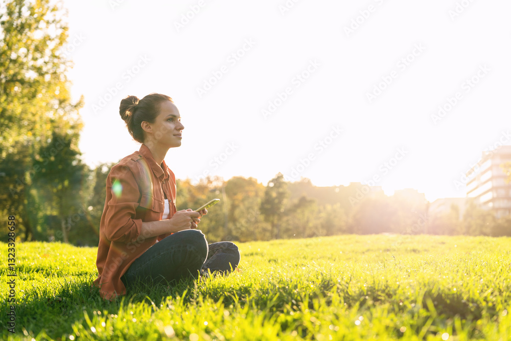 Woman sitting on the green grass outdoors holding mobile phone and looking into the distance 