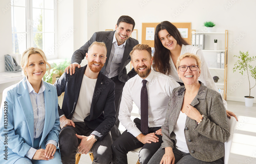 Group portrait of cheerful people colleagues men and women looking at camera and smiling indoors. Company employees in formal clothes sitting on workplace in office. Teamwork and business concept. Group portrait of cheerful people colleagues men and women looking at camera and smiling indoors. Company employees in formal clothes sitting on workplace in office. Teamwork and business concept.