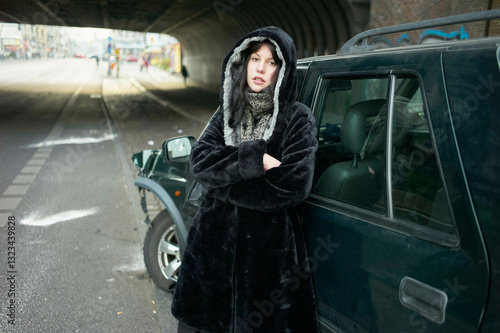 Woman in a black coat leans against a car just after an accident under a city bridge with a distant figure walking. Berlin, Germany