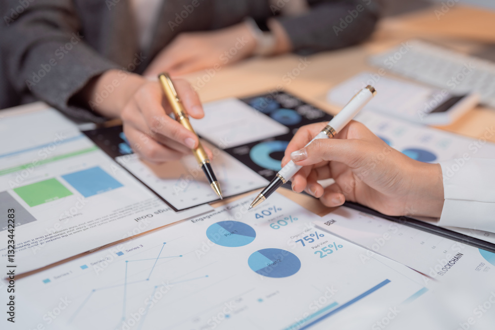 Two businesswomen are collaborating on analyzing financial charts and graphs, using pens to point at key data points and discuss important figures during a corporate meeting Two businesswomen are collaborating on analyzing financial charts and graphs, using pens to point at key data points and discuss important figures during a corporate meeting