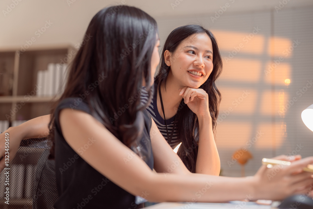 Two young businesswomen are discussing a project sitting at a desk in a modern office, collaborating and sharing ideas in a productive work environment Two young businesswomen are discussing a project sitting at a desk in a modern office, collaborating and sharing ideas in a productive work environment