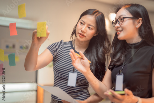 Two young businesswomen collaborating on a project, using sticky notes on a glass wall during a brainstorming session, generating innovative ideas and strategies together