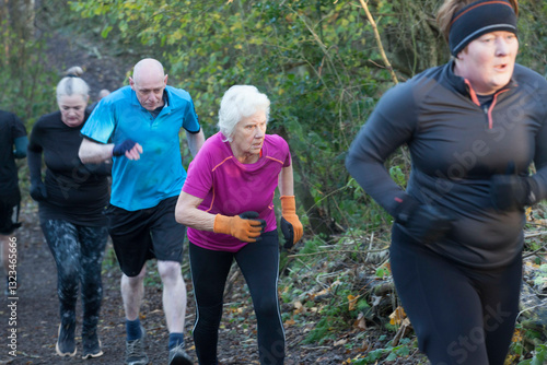 A group of people jogging uphill on a forest trail, dressed in athletic clothing appropriate for cool weather. Sale Water Park, Manchester, UK