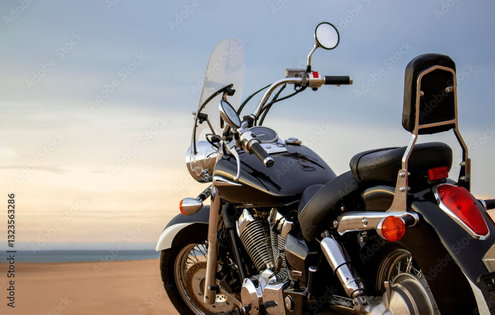 Naklejka premium A lone motorcycle stands on the sandy surface of the Namib Desert against a dramatic cloudy sky. The sunset lighting highlights the contrast between nature and machine.