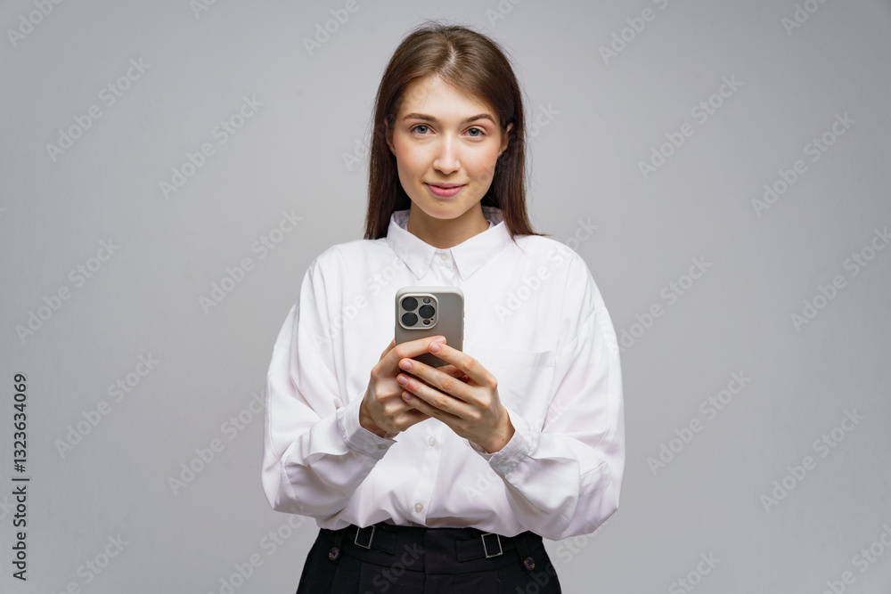 Young woman in white shirt holds smartphone while looking confidently at the camera in a neutral background Young woman in white shirt holds smartphone while looking confidently at the camera in a neutral background