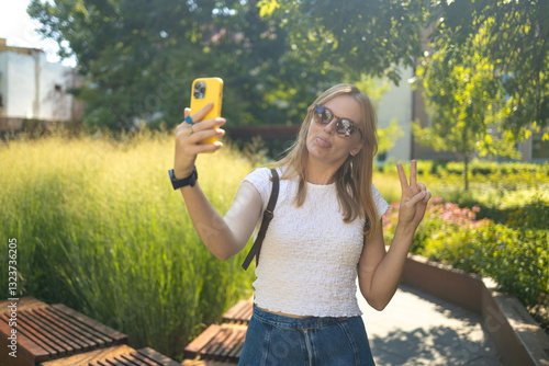Stylish smiling 30s lady taking selfie outdoors in city park, happy young female capturing self-portrait while enjoying walk and good day, resting outside