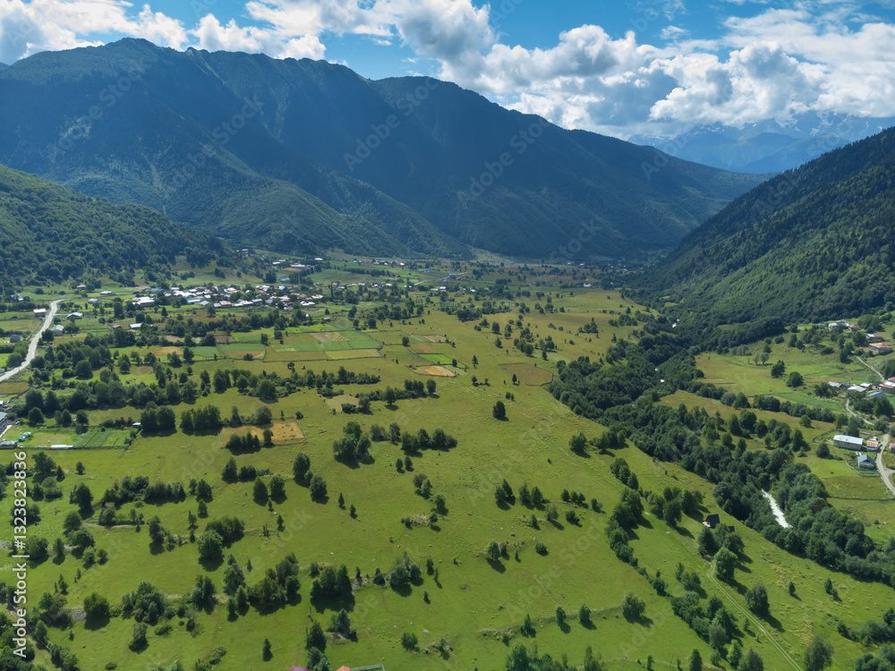 Offering an aerial view of a vibrant green valley surrounded by majestic mountains, featuring a charming village under the warm glow of natural sunlight