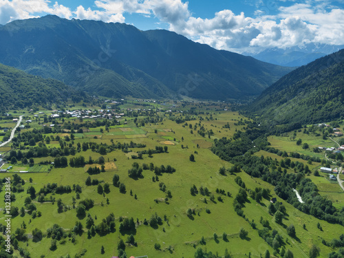 Offering an aerial view of a vibrant green valley surrounded by majestic mountains, featuring a charming village under the warm glow of natural sunlight