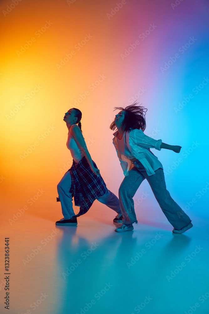 Naklejka premium Dynamic hip hop performance by two young women in casual clothes against gradient studio background in neon light. Concept of movement, breakdance, music culture, artistic expression, youthful energy.