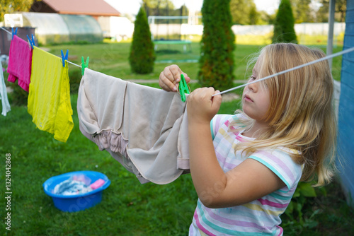 Fotografia Wearing a cheerful summer dress, a six-year-old girl stretches her small arms to hang a damp shirt, her face full of focus