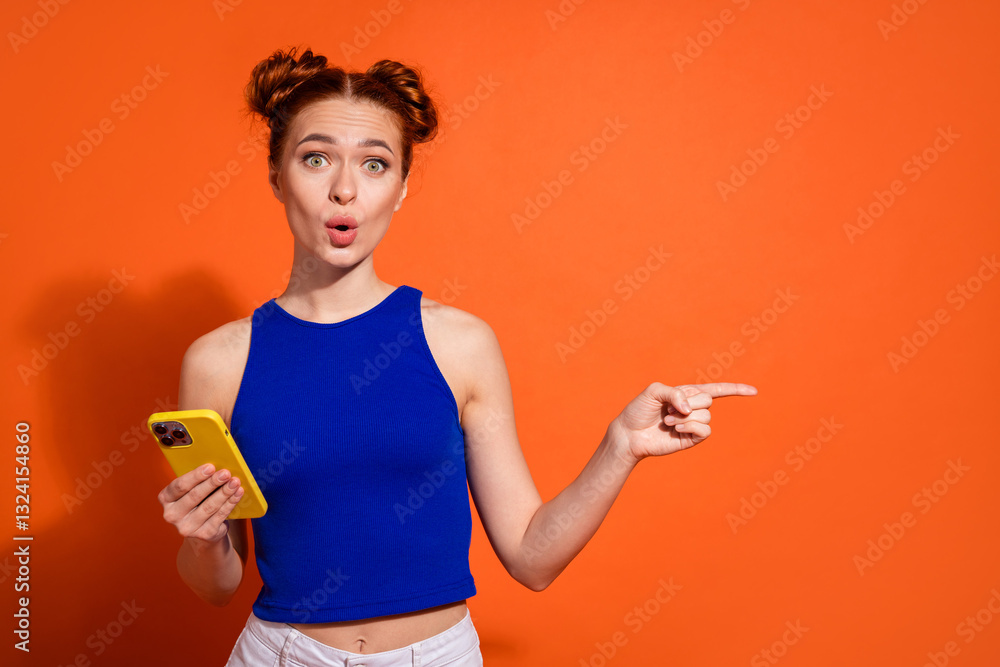 Surprised Young Woman Holding Smartphone and Pointing to the Side on Vibrant Orange Background Surprised Young Woman Holding Smartphone and Pointing to the Side on Vibrant Orange Background