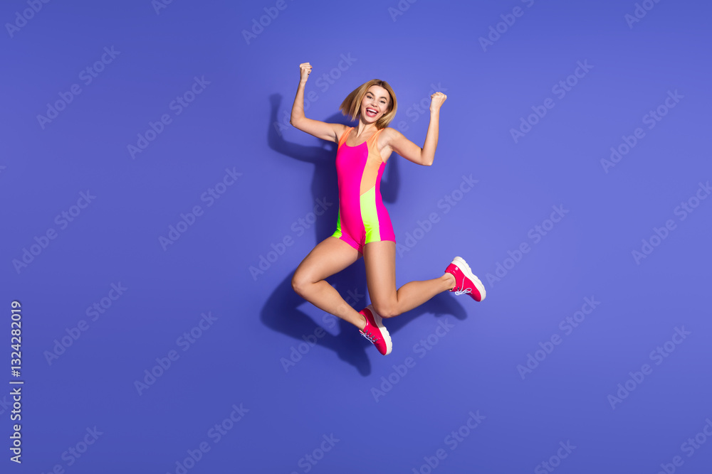 Obraz premium Joyful young woman in vibrant sportswear jumping against a purple backdrop, expressing happiness, fitness, and stylish enthusiasm