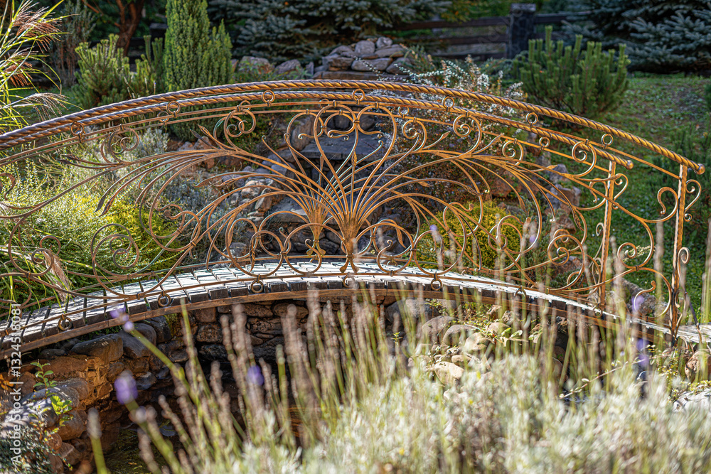 A beautifully crafted wrought iron bridge with intricate golden detailing spans a small garden stream, surrounded by lush greenery and bathed in warm sunlight.