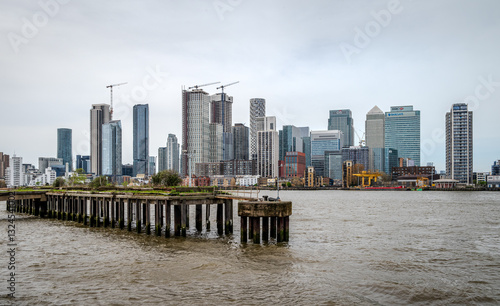 An old pier stands in the Thames River, with the buildings of East London lining the distant bank under an overcast sky