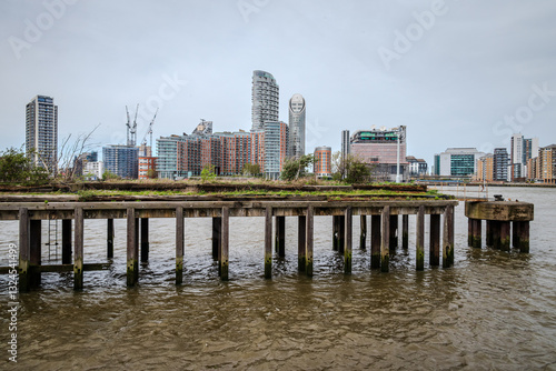 An old pier stands in the Thames River, with the buildings of East London lining the distant bank under an overcast sky