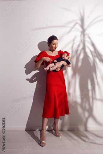 Obraz na płótnie portrait a woman in a red dress holds a child at home on a white background with a shadow from a bush w salonie
